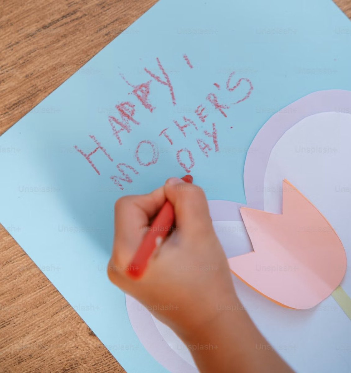 Child's hand writing 'Happy Mother's Day' in red crayon on a blue card with a paper tulip