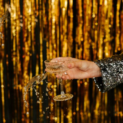 Main en robe pailletée tenant une coupe de champagne devant un rideau doré scintillant